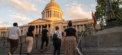 The Washington State Capitol dome in Olympia rises against a warm evening sky, with a small pride flag visible near the entrance steps.