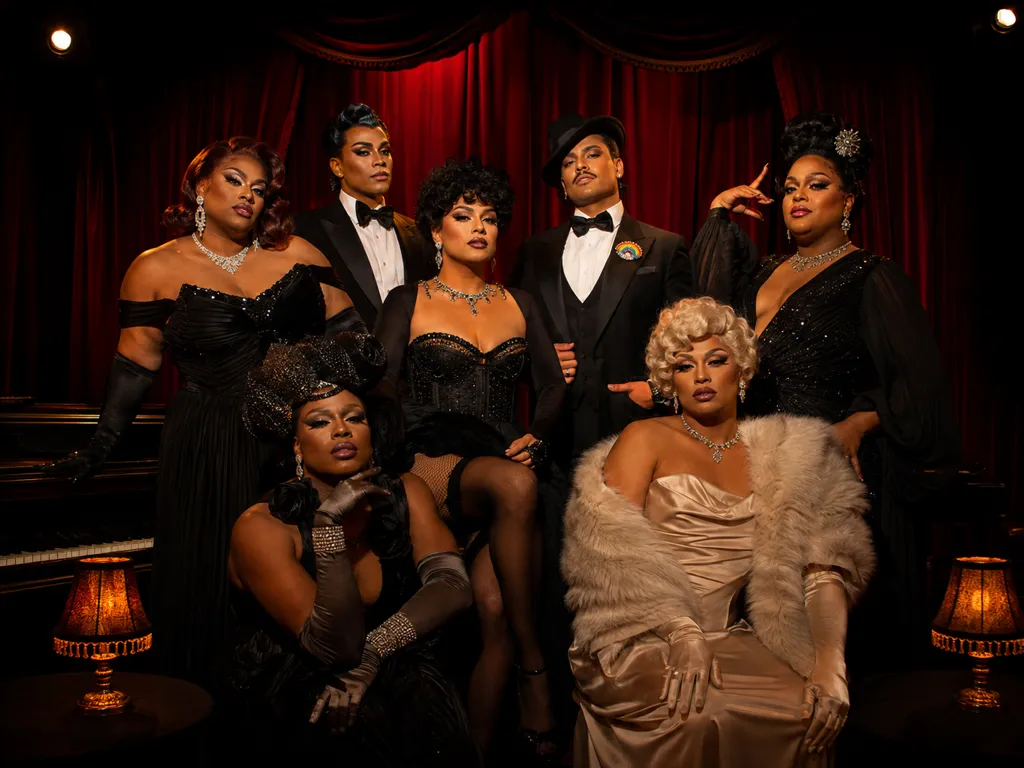 A group of drag performers in glamorous black-tie evening wear poses dramatically before a red velvet curtain, one wearing a rainbow lapel pin.