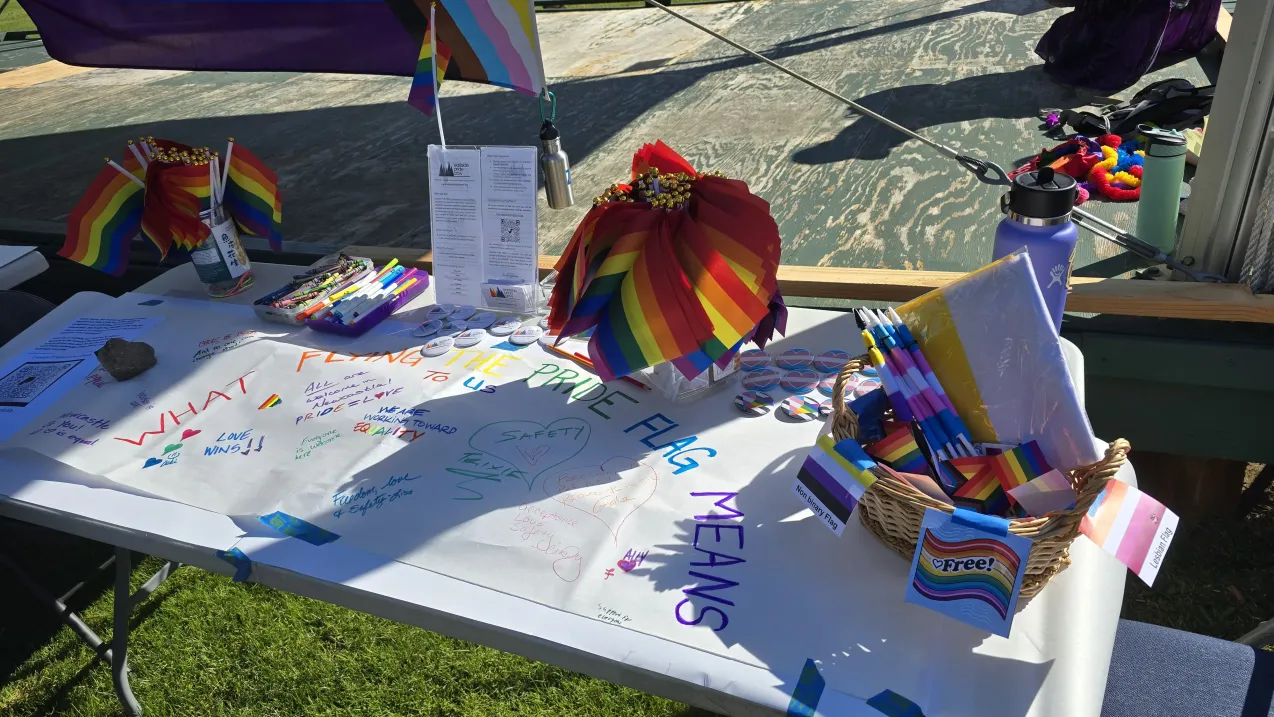 A table at a Pride event covered with rainbow flags, markers, and messages about the meaning of the Pride flag.