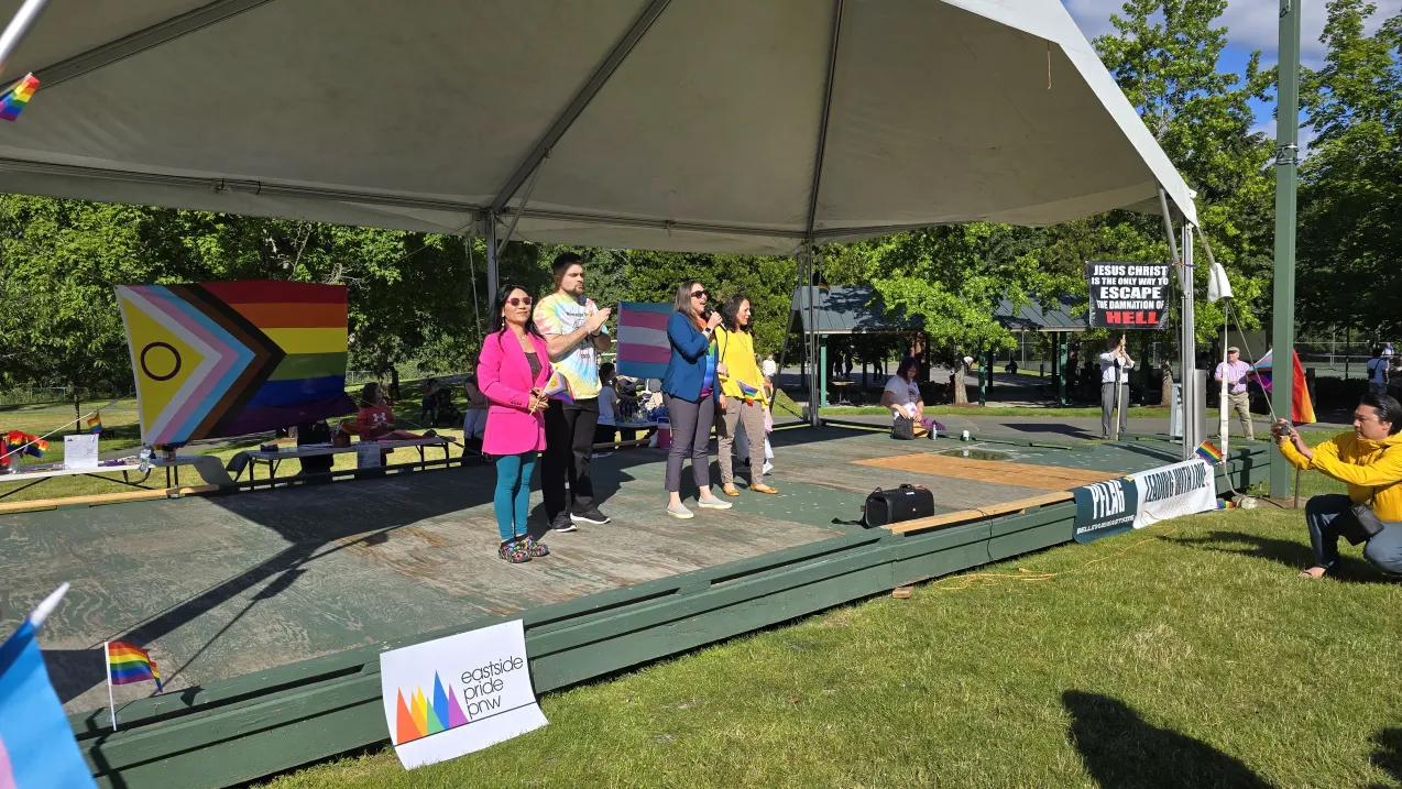 A group of people standing on a stage at a Pride event, with Pride flags displayed and a protestor holding a religious sign in the background.