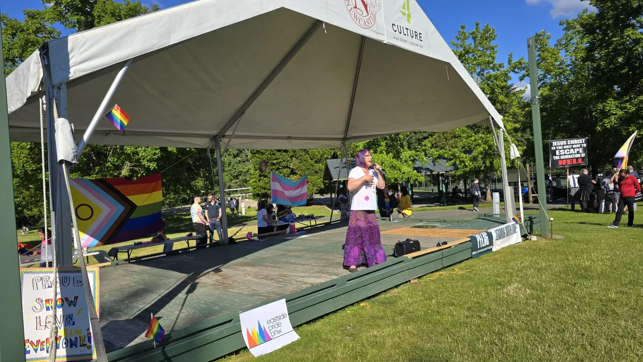 A speaker on stage at a Pride event, with Pride and Transgender flags displayed in the background, while a protestor with a religious sign stands nearby.