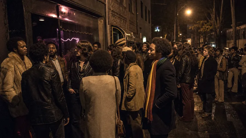 A crowd gathers on a narrow city street outside a lit bar at night in a scene evoking the 1969 Stonewall uprising.